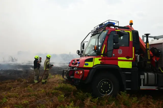 A fire service off-road vehicle on the heathland. There are firefighters aiming a hose at the ground.