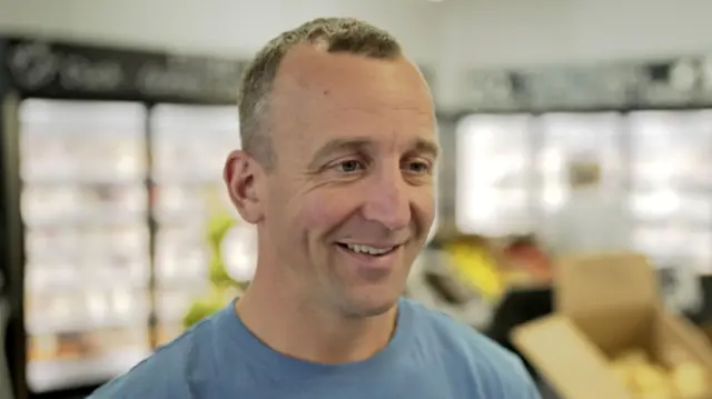 A man looking toward the right hand side of the image who is smiling as he stands inside a farm shop. He has short dark hair and is wearing a blue T-shirt.