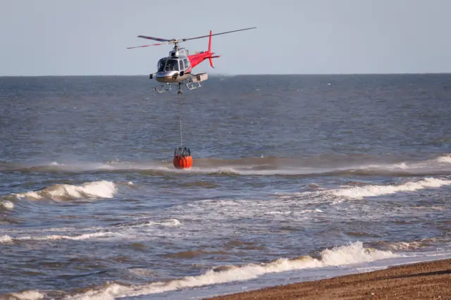 A red and silver helicopter hovering over the North Sea at Dunwich, with its nose slightly down. It is trailing a large red bucket into the sea, just above the waves.