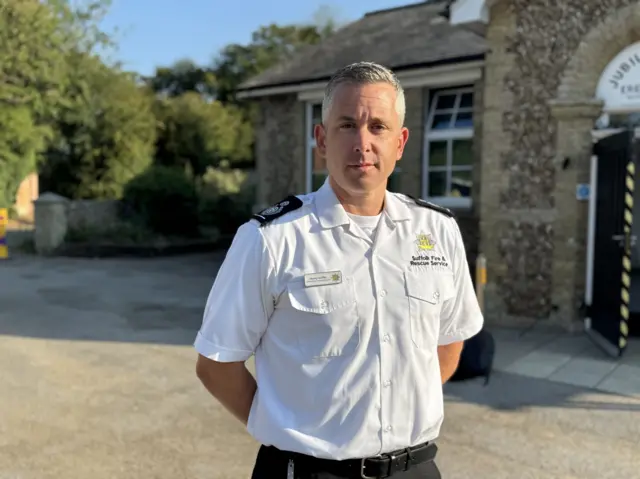 Henry Griffin stands in front of a village hall. He is in a white fire service shirt and black trousers and his arms are crossed behind his back.