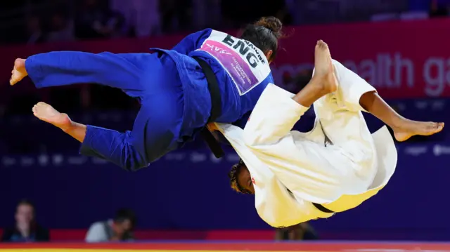 Lele Nairne, wearing a blue robe, flips over Frema Agyei, wearing white, to win bronze by ippon