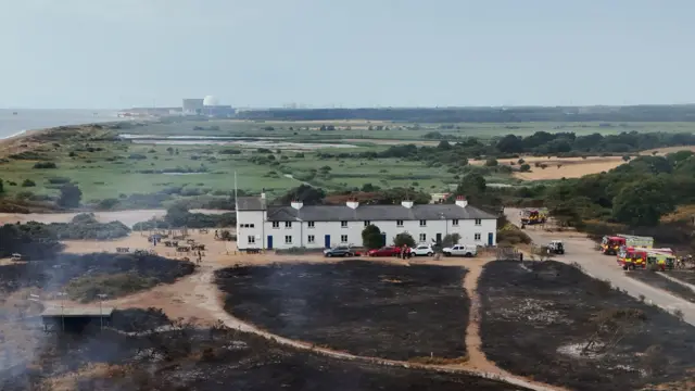 A white row of terraced cottages, with black scorched earth in the foreground. The white dome of a power station is in the background beside the sea.