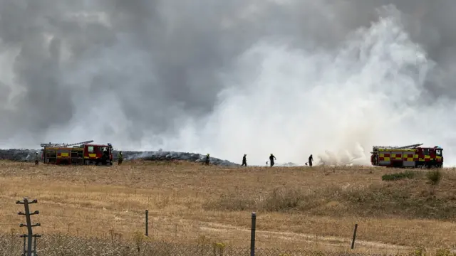 Two yellow and red fire engines are parked on dry brown grass, and behind them is white billowing smoke. A metal fence is in the foreground.