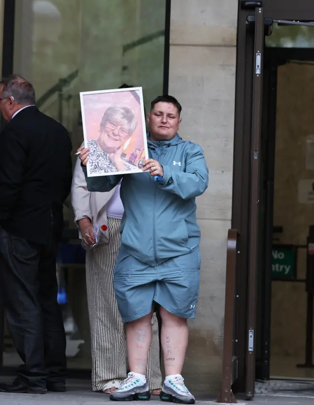 A family member holding a framed photo of a woman with short blonde hair and glasses, they are standing on the steps of Hull Crown Court
