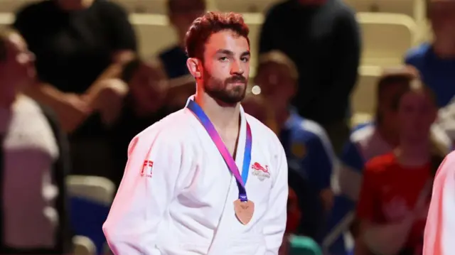 Micahel Fryer, wearing a white robe, stands on the podium with his bronze medal around nhis neck