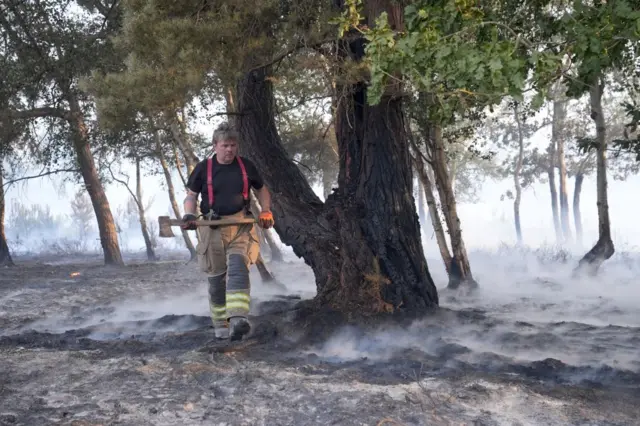 A firefighter carrying an axe walks through the smouldering remains of a wildfire at Dunwich Heath. Smoke is still coming up from the scorched earth.