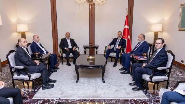 Six men in suits are shown sitting in chairs around a small coffee table, with a Turkish flag in the background.