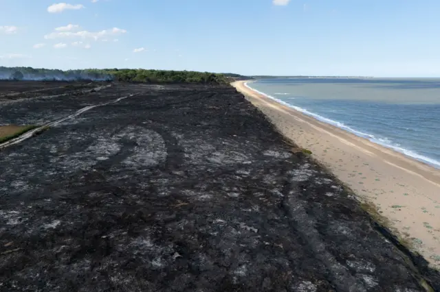 Scorched black earth for at least a mile is on the left of the frame. To the right is a beach and the blue sea.