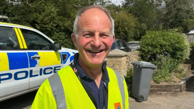A man smiles at the camera as he stands outside a village hall on a sunny day. He is standing in front of a police car. He has short grey hair and is wearing a hi-vis jacket with a blue polo top underneath.
