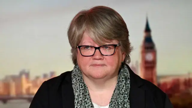 A woman sitting in a TV studio looking away from the camera. She is not smiling. She has short blonde hair and is wearing a black jacket with a grey scarf with black and white spots on it,with a white T-shirt underneath.