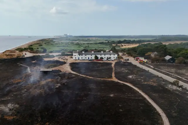 A row of white cottages and cars parked outside. In front of it, in the foreground, is black scorched earth. Behind are green fields, and the sea to the left.