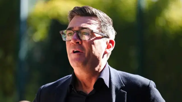 Andy Burnham is pictured speaking against a background of greenery, with sunlight falling across his face. He is looking off to the left of camera.