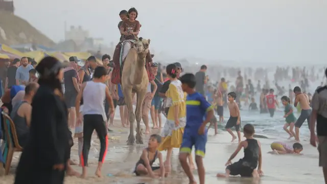 A large crowd of people is shown in and around the water at a beach, with several young people riding a camel and laughing.