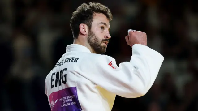 Michael Fryer, wearing a white judo robe with his name and ENG on the back, pumps his right fist in the air to celebrate winning bronze