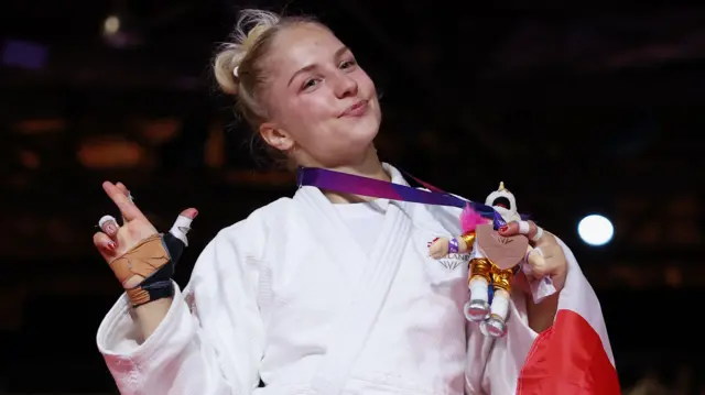 Tatum Keen, wearing a white judo robe, smiles and holds up her bronze medal and mascot while also clutching an England flag