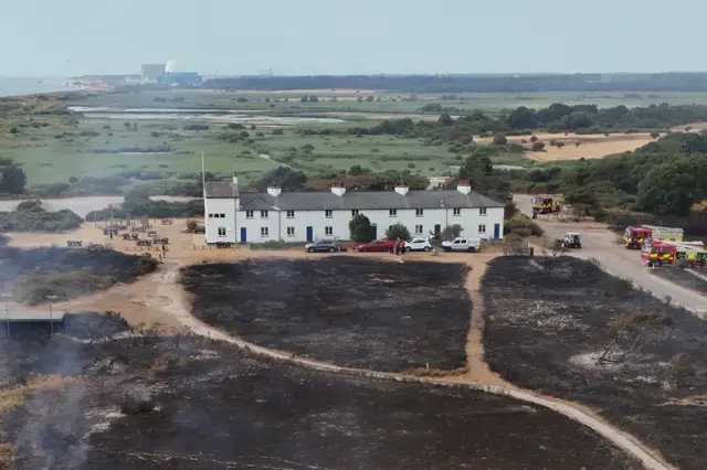 A heathland area that has been burnt during a wildfire. A row of white cottages sit in the middle of the field, untouched by the fire. Fire crews and vehicles are parked nearby on a road. Firefighters walk around the scene. Some smoke billows upwards around the area.