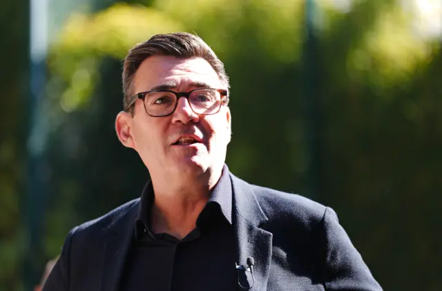 A close-up of Andy Burnham, showing his head and shoulders. He has dark hair, is wearing dark-framed glasses and a black jacket over a black polo shirt.