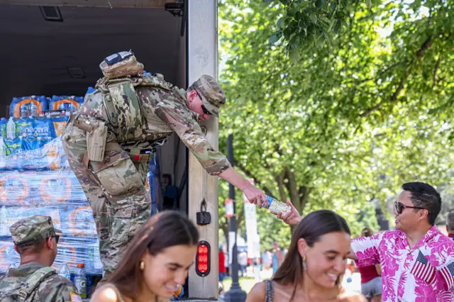 A members of the National Guard wearing a camouflage uniform hands a water bottles to a man in a pink, patterned shirt with a small US flag slotted in his pocket