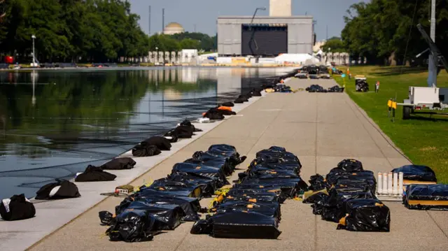 Several black bags have placed in rows on stone paving on the right side walkway of the Reflecting Pool in Washington. There is also a long line of black bags, which seem to be made from a different material, placed right on the edge of the Reflecting Pool. The base of the Washington Monument is seen in the distance behind a stage.