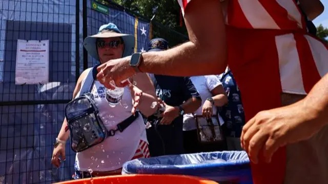 A woman wearing a rucksack on her front is handed a bottle of war that had been being kept cool in a bucket of ice