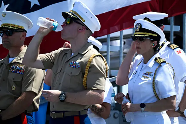 A soldier drinks a bottle of water. He is wearing sunglasses and a white hat with his military outfit