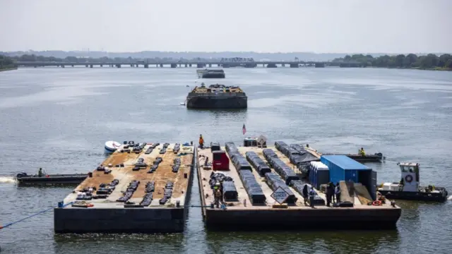 Three pontoons pictured floating in a line in the Potomac River. They are each carrying dozens of black bags, which have been arranged in rows and supposedly contain fireworks. At the nearest pontoon, two small boats are pushing it from the right, while another small boat is pushing from the left. A bridge can be seen in the far distance crossing the river.