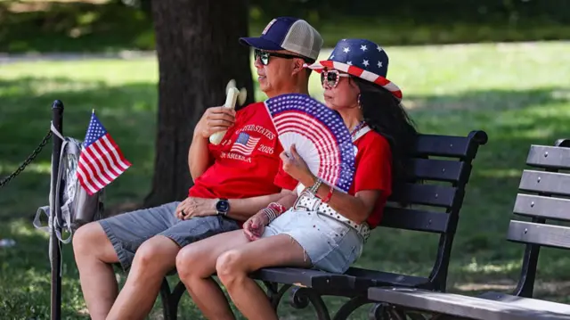 A man and a woman covered head to toe in American-themed clothing sit on a park bench in the shade. The man is holding an electric fan pointed towards his face, while the woman uses a paper fan on herself. They are both wearing sunglasses and hats to combat the hot weather.