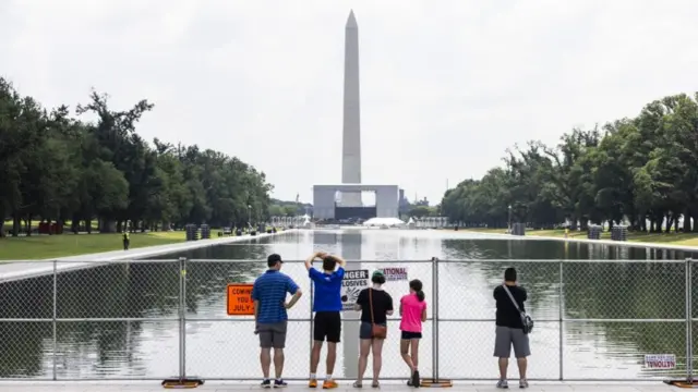 A group of five people are stood next to fencing that surrounds the Lincoln Memorial Reflecting Pool. One person to the right appears to be taking a picture of the pool, while what appears to be a family of four stand to the left. The Washington monument is seen in the distance on the other side of the reflecting pool.