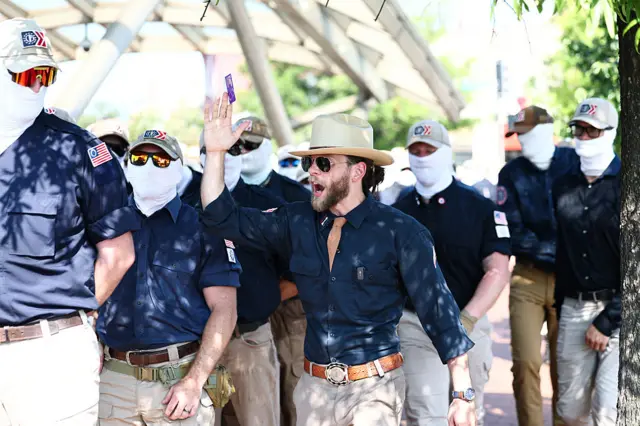 Patriot Front founder Thomas Rousseau wearing a large brimmed beige hat, partially unbottoned blue shirt and beige khakis surrounded by members of the group in matching outfits