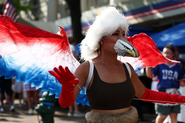 A woman dressed as an eagle with red, white and blue wings. She has a puppet of an eagle's head strapped over her nose and mouth