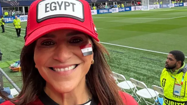 A woman in a red football jersey tales a selfie while wearing a hat that says Egypt while posing in front of a large football stadirum