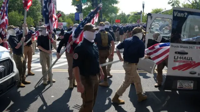 Members of the white supremacist group Patriot Front loads US flags into a van in Washington. Behind them is a larger group of men who appear to be lined up in unison. They are all wearing white balaclavas, sunglasses and hats, with dark blue shirts and beige trousers.