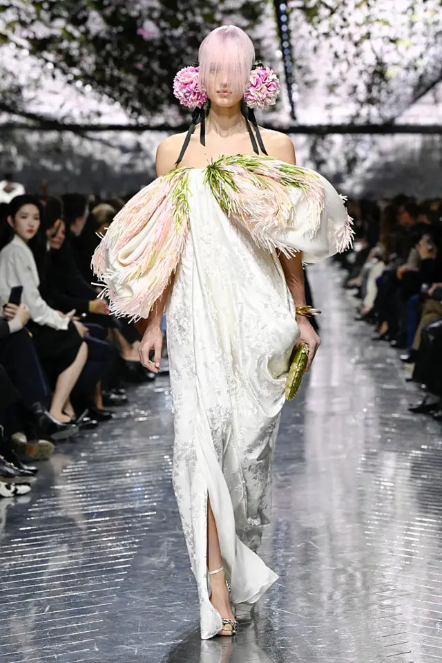 A model walks the runway wearing a drapey white dress with oblong shapes on the sides