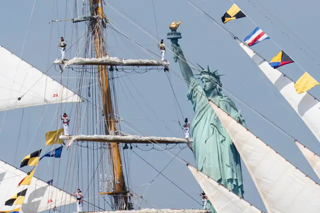 Six sailors stand at the end of a mast with its sails rolled in. The Statue of Liberty can be seen clearly behind the boat