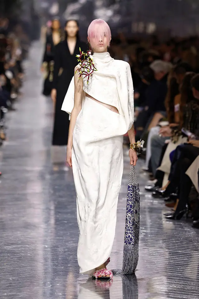 A model walks the runway wearing a long white dress that resembles a Grecian robe