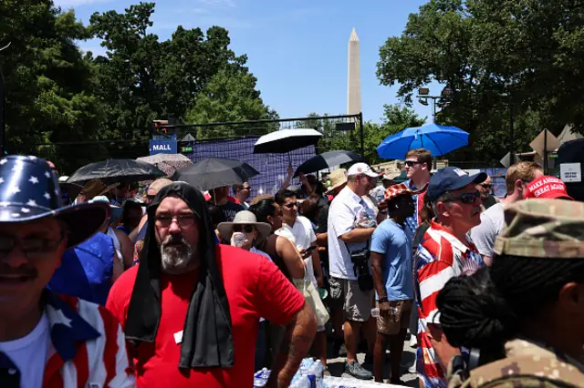 A crowd of people, some with umbrellas, others with cowboy hats, stand around. The Washington Memorial can be seen in the background