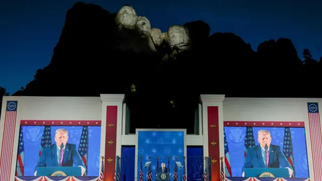 US President Donald Trump speaks between two big screens in front of Mount Rushmore. The stone faces of the monument are lit up in the dark