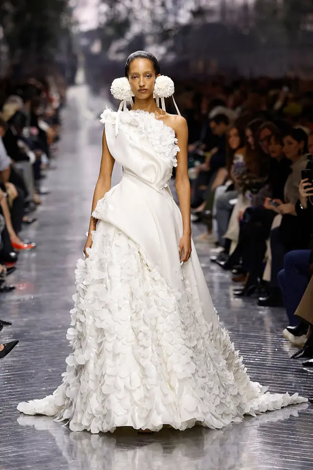 A model walks the runway wearing a long white dress with frilly accents.