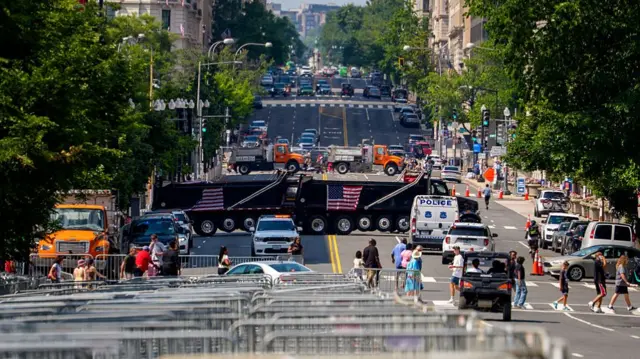 A view looking down 14th Street NW along the National Mall. Several large trucks are spread across the road lengthways at two different points along the street. Rows of metal barriers are seen at the fore front as people walk along a zebra crossing behind them. Rows of trees line each side of the road, next to blocks of large high rise buildings.