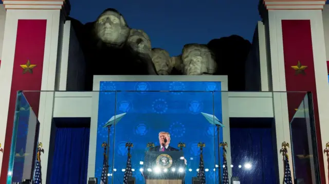 Donald Trump delivers a speech on a stage in front of Mt Rushmore. The four faces of the sculpture are lit up against the dark