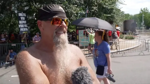 Shirtless man with orange-tinted wide sunglasses speaks into a microphone.