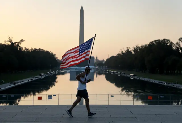 Man carries an American flag during a morning run past the Reflecting Pool as the city prepares for July 4th festivities on July 03, 2026, in Washington, DC.