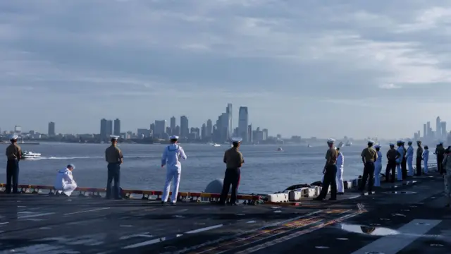 Navy and marine officers wait on board the USS Kearsarge ahead of the Sail4th 250 in New York Bay, the skyline of the Big Apple visible in the distance