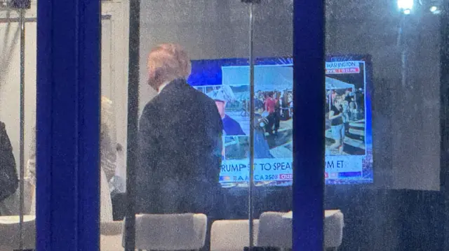 A man can be seen through a rectangular window, watching news coverage on a large television screen.
