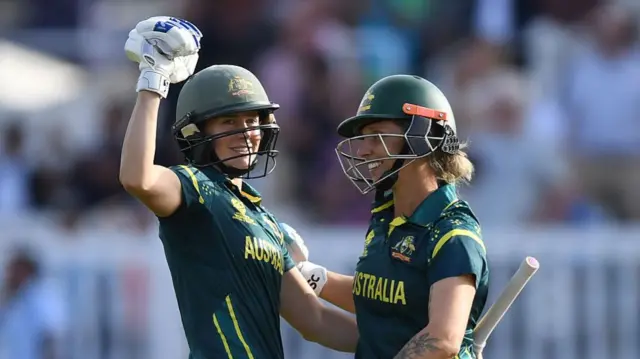 Ellyse Perry (left), wearing a dark green Australia shirt and helmet, embraces Ash Gardner and raises a gloved left fist