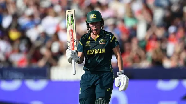 Beth Mooney, wearing a dark green Australia kit and cap, raises her bat in her right hand to celebrate a half-century