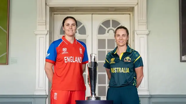 Nat Sciver-Brunt (wearing red) and Sophie Molineux (wearing green) standing either side of the silver t20 World cup trophy
