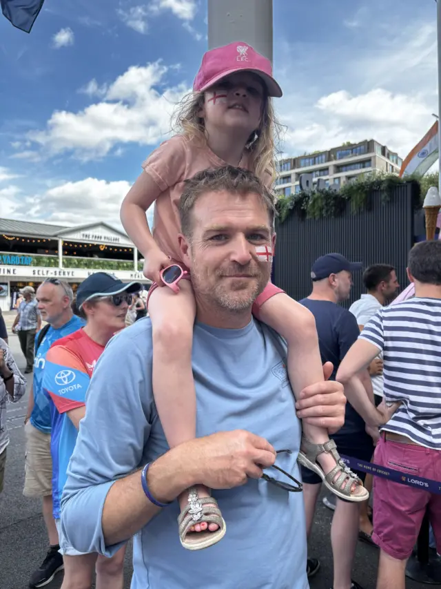 Man in a pale blue jumper with his daughter, wearing a pink top and cap, on his shoulders
