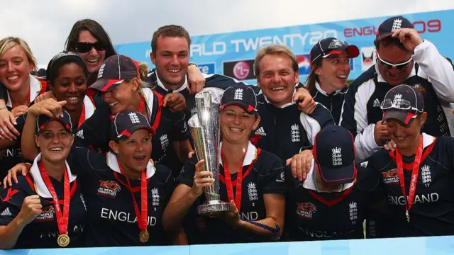 Charlotte Edwards, wearing a dark blue England top and cap, wears a medal on a red ribbon around her neck and holds the silver T20 World Cup trophy with her teammates gathered around her