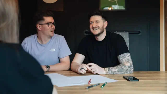 Andy Golpys, on the right, smiles as he sits with colleagues. He is wearing a black t-shirt and has tattoos on his arm.